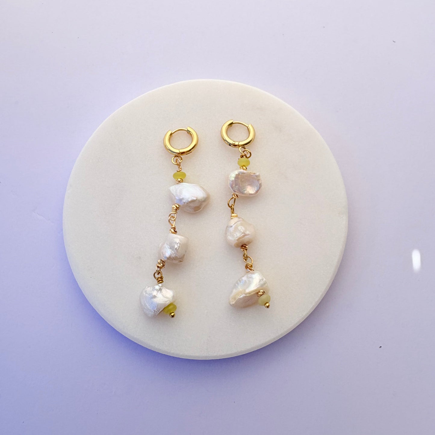 Two pearl and gold bracelet sets on a round white stone surface with a light purple background.